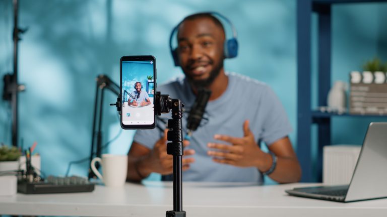 A man wearing headphones speaks into a microphone while recording himself on a smartphone mounted on a tripod at a desk with a laptop and coffee mug.