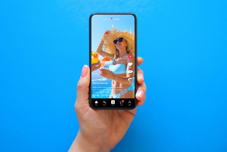 TikTok Experiments With Letting Users See Less AI Slop A hand holds a smartphone showing a TikTok video of a woman in a blue bikini, straw hat, and sunglasses, holding a drink against a bright blue background.