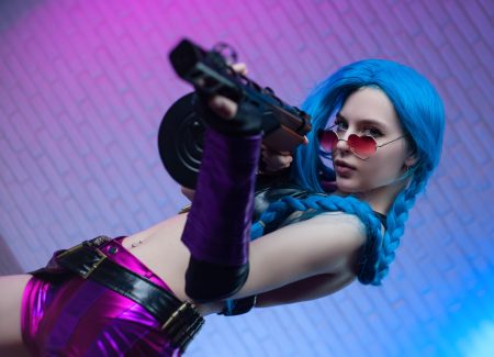 Person with long blue braided hair, wearing pink sunglasses and shiny purple outfit, holds a large gun and poses in front of a brick wall with pink and blue lighting.