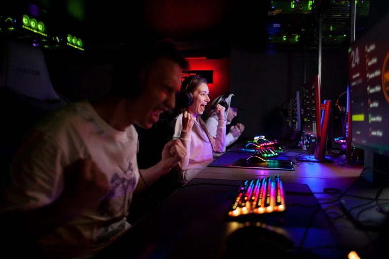 Two people at gaming computers wearing headsets react with excitement, clenching their fists, illuminated by colorful keyboard and monitor lights in a dark room.