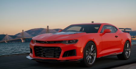 A red sports car driving on a road near water, with a suspension bridge and mountains in the background at sunset.