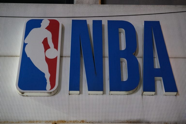 The NBA’s Most Exciting Young Stars Right Now The image shows the NBA logo featuring a white silhouette of a basketball player alongside large blue letters spelling "NBA" on a perforated white background.