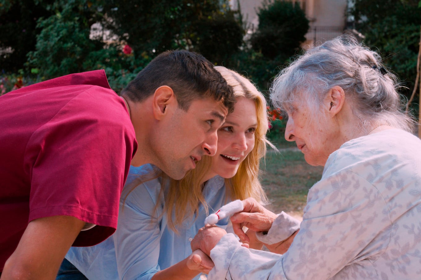 A young man and woman interact warmly with an elderly woman outdoors, holding her hands and smiling at her.