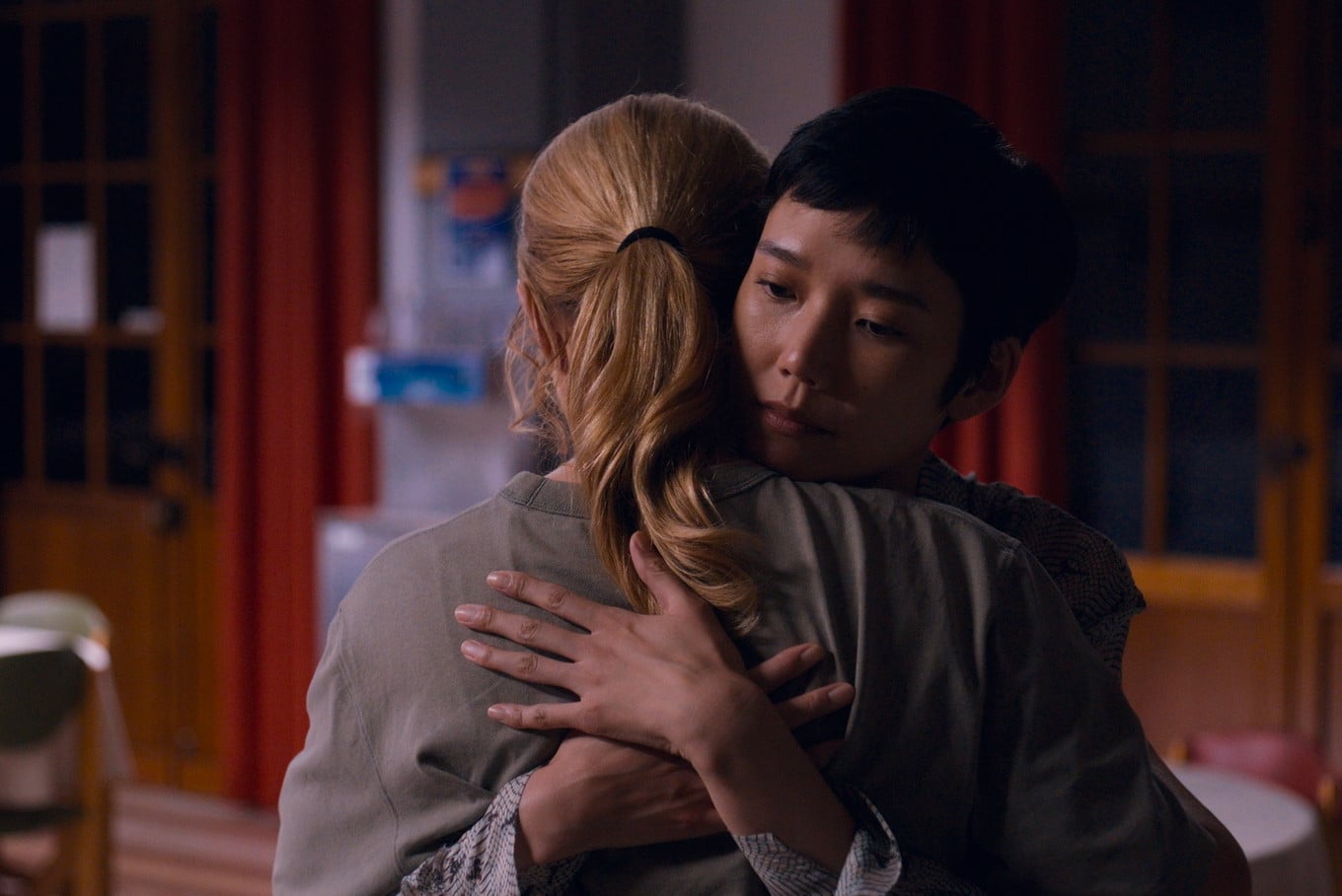 Two people are embracing in a dimly lit room, with one facing the camera and the other facing away. The background shows chairs, a table, and windows with red curtains.