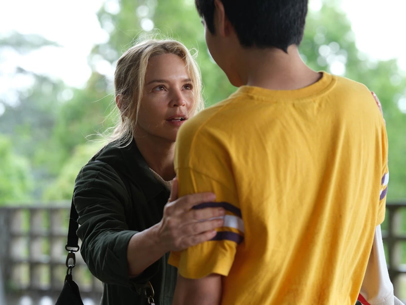 A woman with blonde hair holds the arm of a person in a yellow shirt, looking at them intently outdoors with greenery in the background.