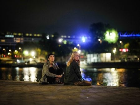 Two people sit on a riverside pavement at night, looking up. City lights and reflections shimmer on the water in the background.