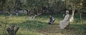 ‘Black Rabbit, White Rabbit’ Blu-Ray Review – Shahram Mokri Delivers His Latest Engaging Oddity A woman in a light dress sits on a chair, while a child crouches beside a basket in a grassy orchard with several fruit trees.