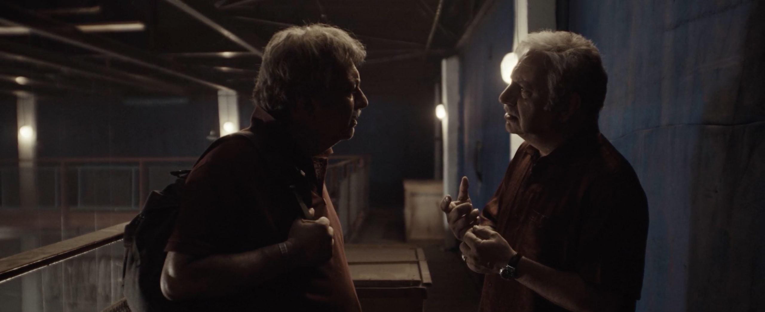 Two older men stand indoors, facing each other and talking; one has a backpack and holds the straps, while the other gestures with his hands.