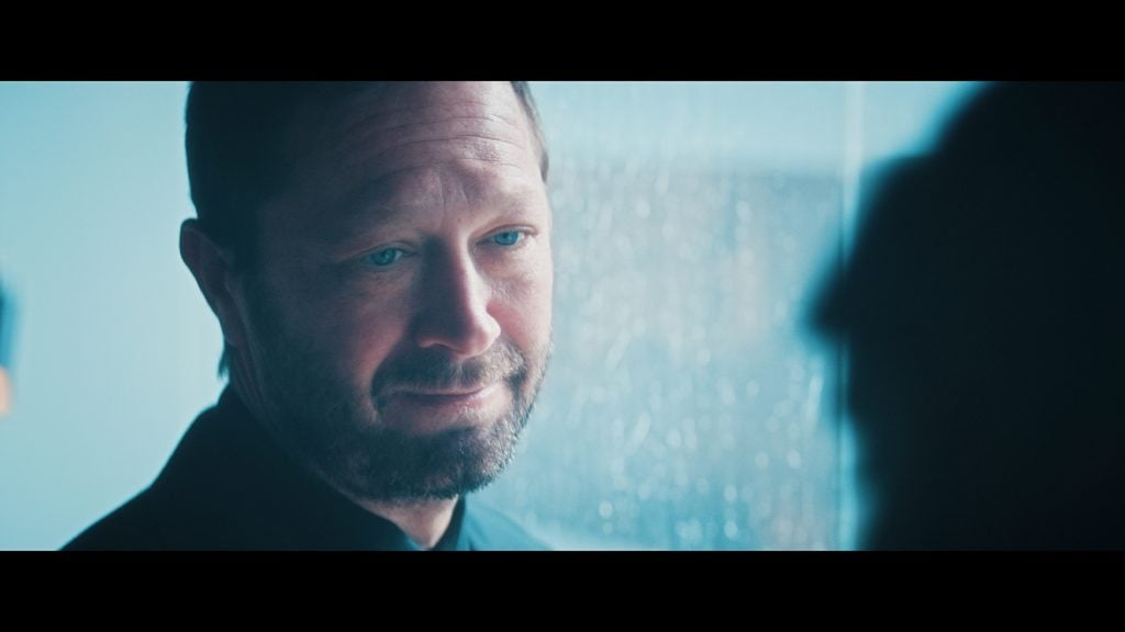 A man with a beard and short hair stands indoors near a rain-streaked window, looking towards someone out of focus in the foreground.