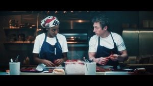 Two chefs in white shirts and blue aprons stand behind a counter, preparing food in a commercial kitchen. They appear to be having a serious conversation.