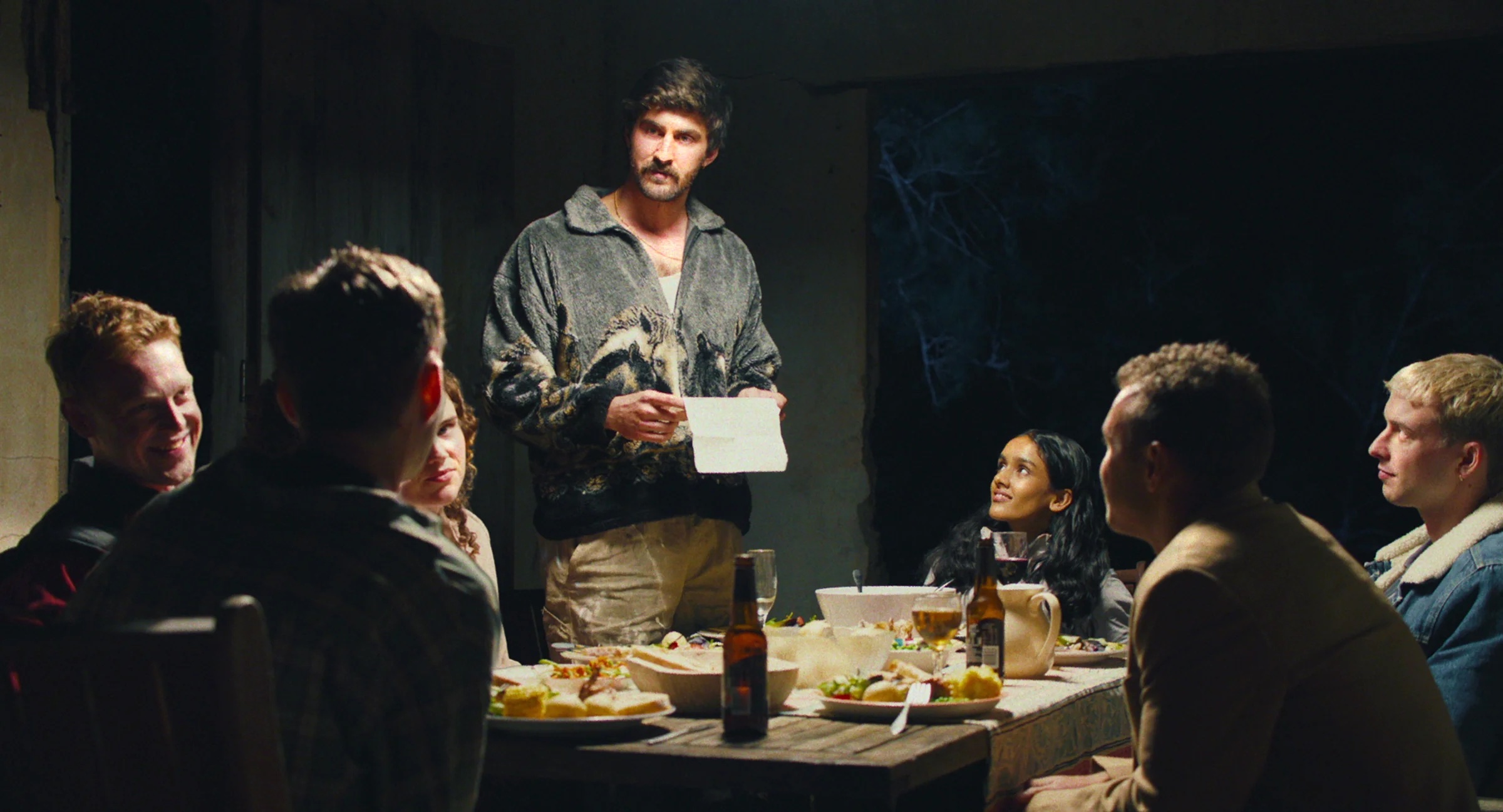A man stands holding a piece of paper, speaking to five seated people around a dinner table with food and drinks in a dimly lit room. A still from Birdeater. 