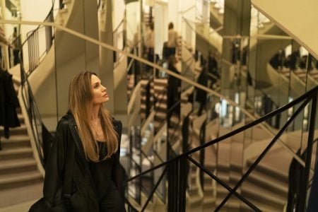 A woman with long hair stands on a staircase surrounded by mirrors, looking upward, with multiple reflections visible in the background.