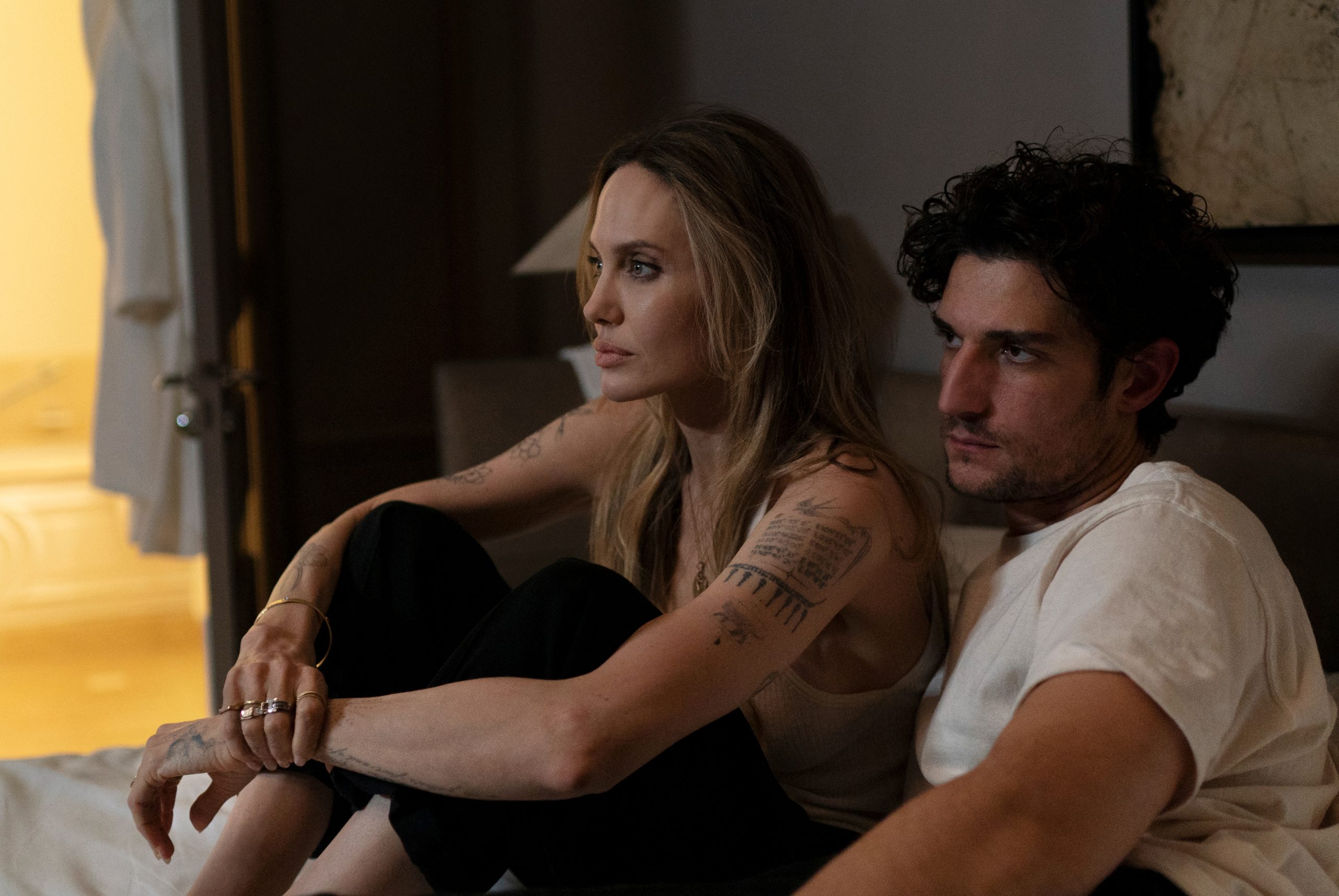 Two people sit closely together on a bed, both looking intently in the same direction. The room is dimly lit, and their expressions are serious.