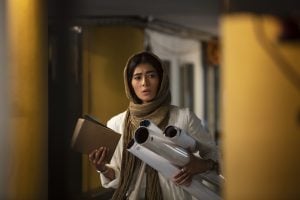 A woman wearing a headscarf holds rolled-up papers and a notebook, standing indoors with a serious expression.