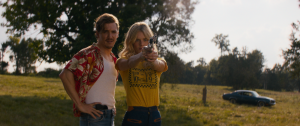 ‘Carolina Caroline’ Review – Weaving And Gallner Electrify In This Road Trip Crime Thriller A man stands beside a woman aiming a handgun in an open grassy field, with trees and a parked car visible in the background.