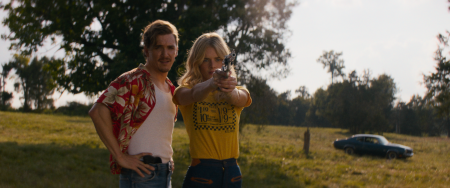 ‘Carolina Caroline’ Review – Weaving And Gallner Electrify In This Road Trip Crime Thriller A man stands beside a woman aiming a handgun in an open grassy field, with trees and a parked car visible in the background.