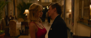 A man and a woman stand closely facing each other in a dimly lit, elegant hallway, with the woman in a red dress and the man in a dark suit.
