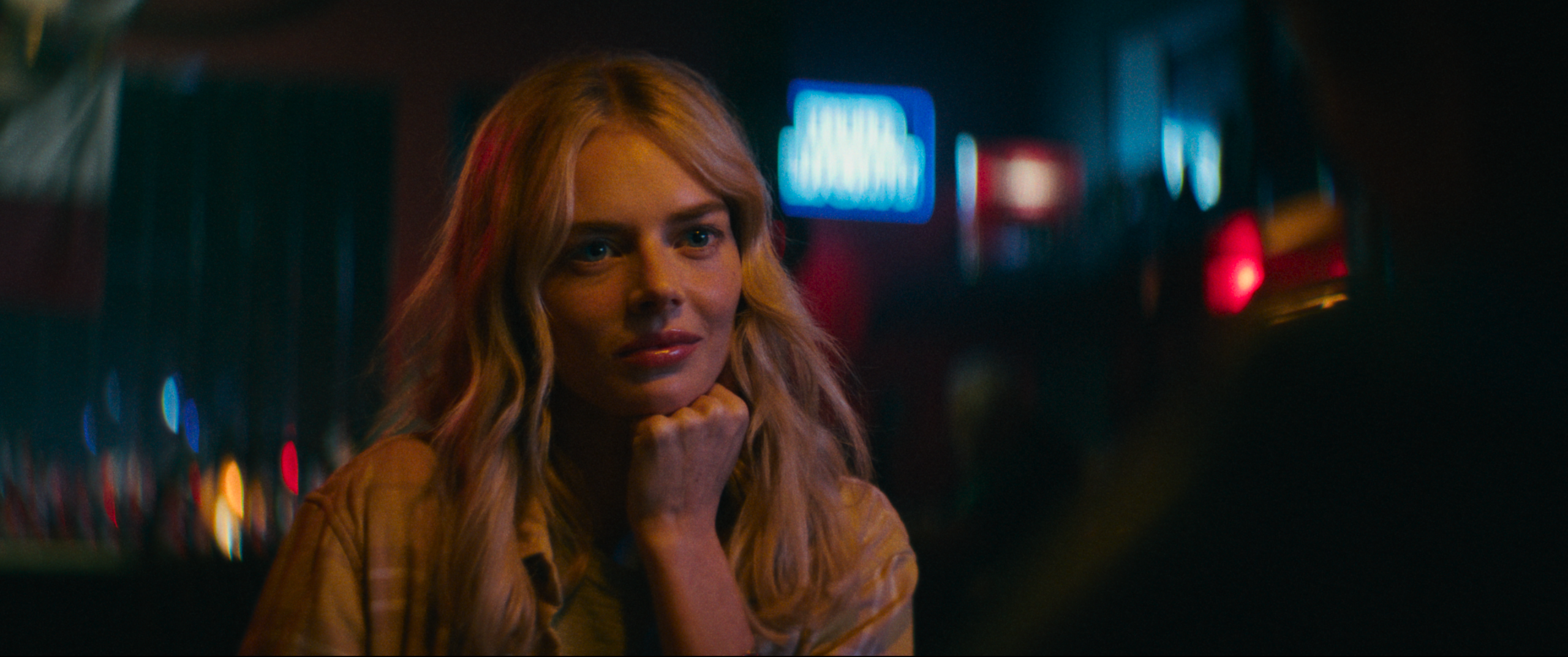 A woman with long blonde hair sits indoors, resting her chin on her hand, looking attentively at someone. The background is dimly lit with colorful lights.
