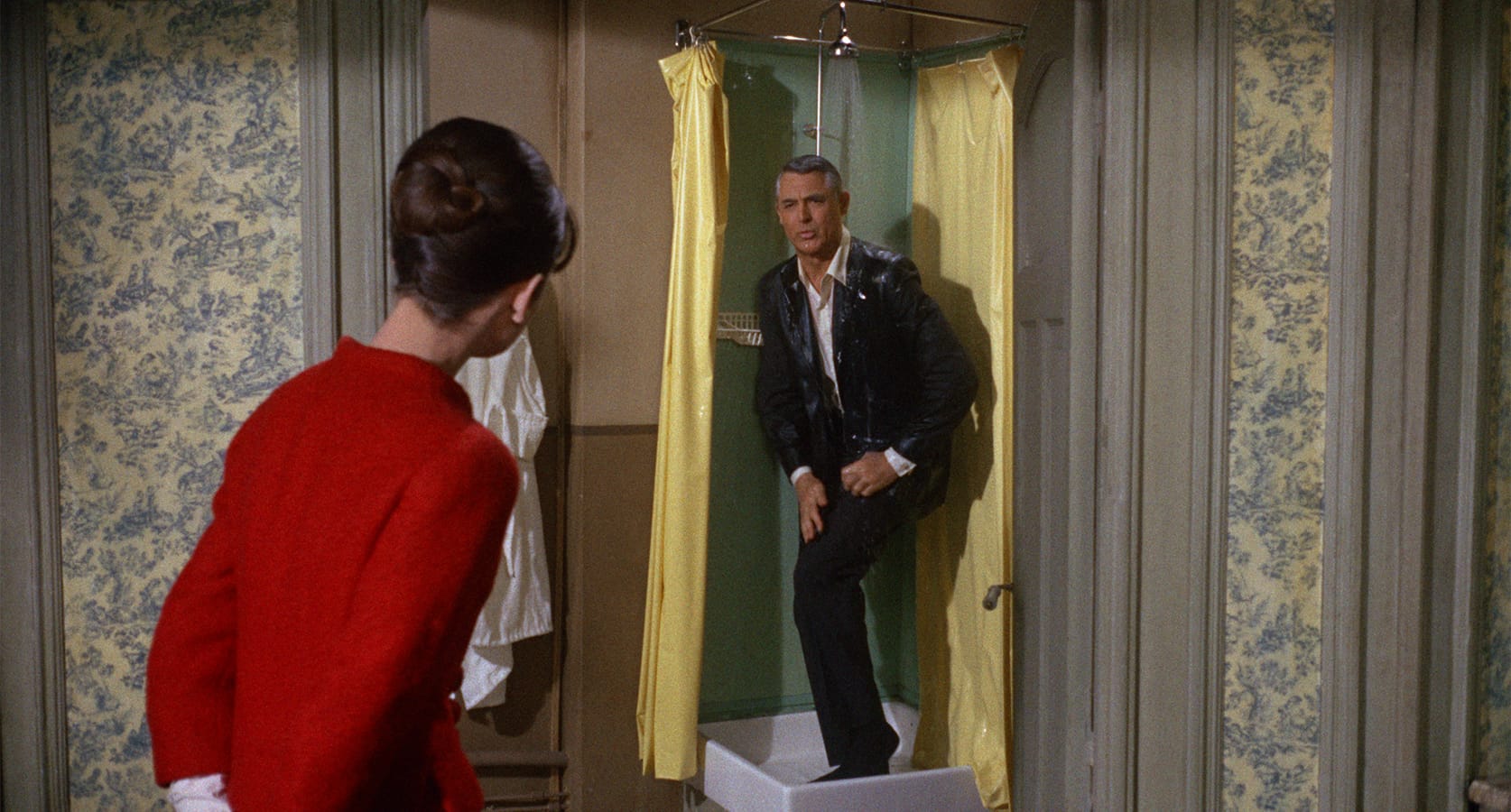 A woman in a red outfit faces a man in a suit standing soaked in a shower with yellow curtains in a vintage-style bathroom.