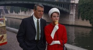 A man in a suit stands beside a woman in a red coat and white hat eating an ice cream cone by a riverside with a bridge in the background.