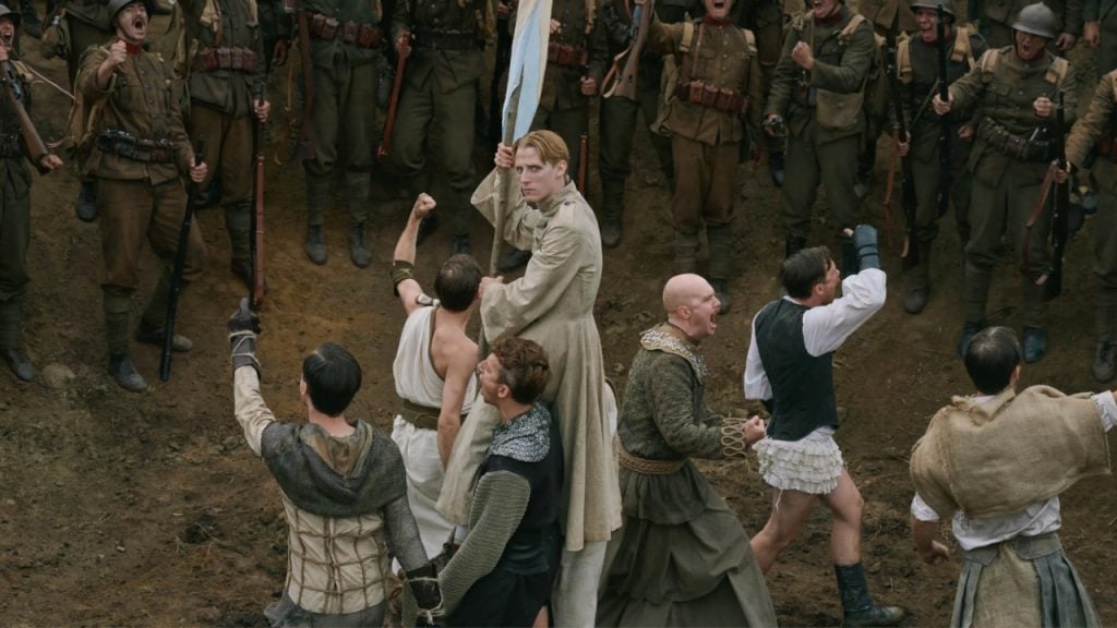 A group of actors in period costumes stand in a circle, raising their fists, with soldiers in uniform cheering in the background.