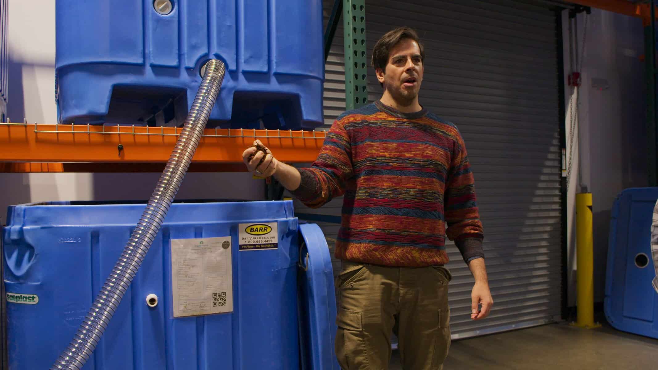 A man in a striped sweater stands indoors holding a hose attached to a large blue storage tank on a metal shelf.