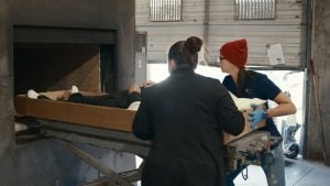 Two people in work attire load a cardboard container with a body inside into a cremation chamber inside a facility.