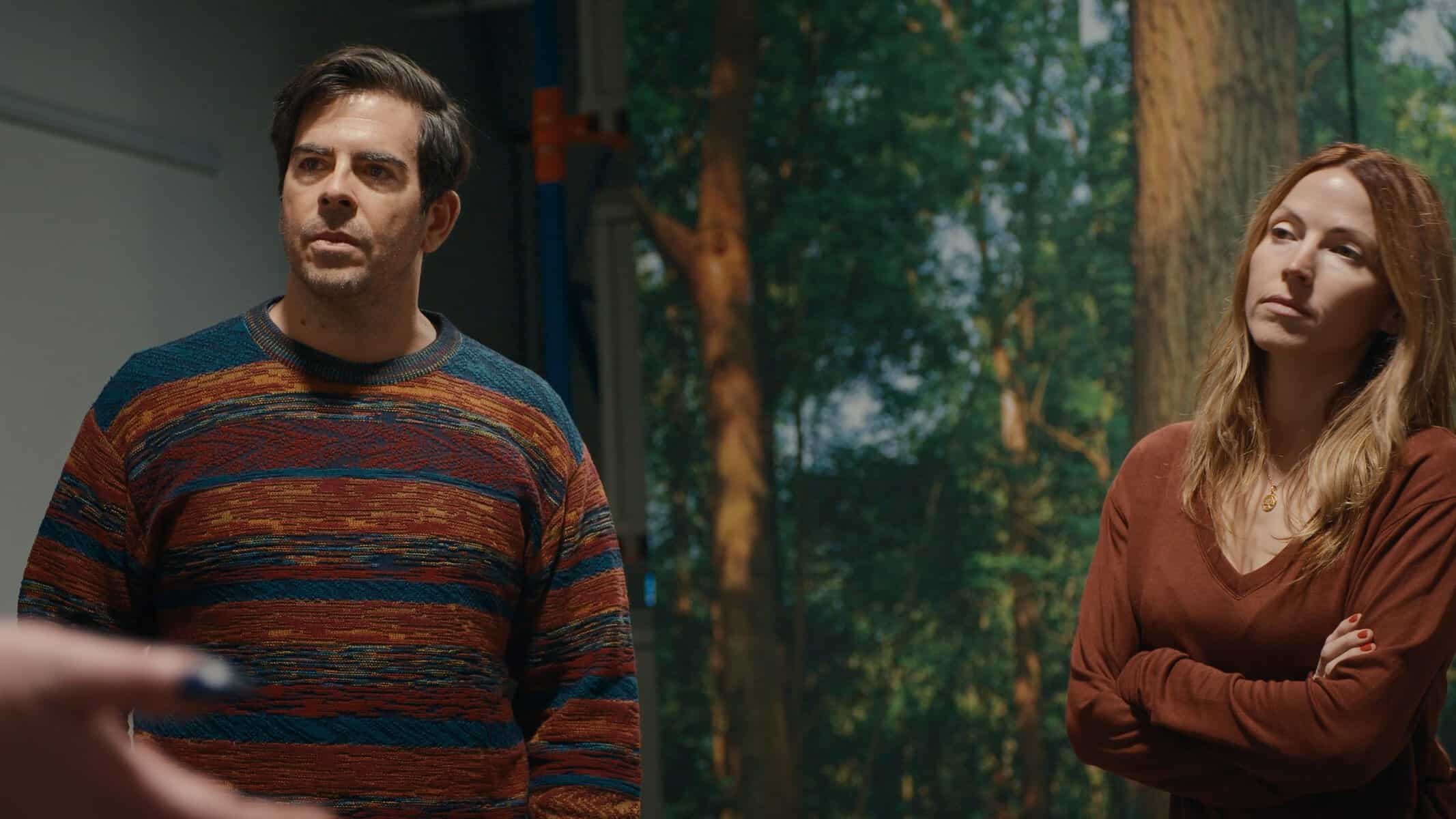 A man in a striped sweater and a woman in a brown top stand indoors, with a forest backdrop visible behind them. Both appear to be listening attentively.