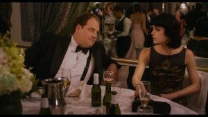 A man in a tuxedo and a woman in a black dress sit at a table with drinks, looking at each other at a formal event with other guests in the background.