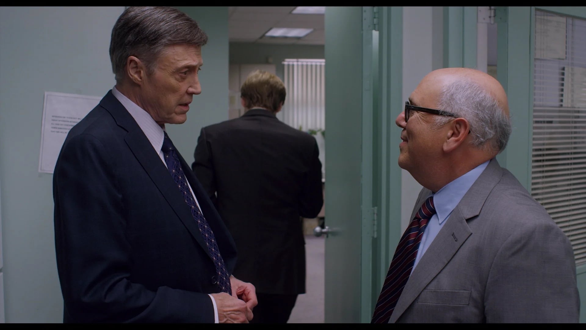 Two men in suits converse in an office hallway while a third man walks away in the background.