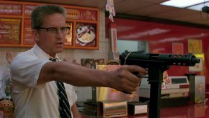 A man in glasses and a white shirt points a gun inside a fast food restaurant, standing in front of the counter with a tense expression.