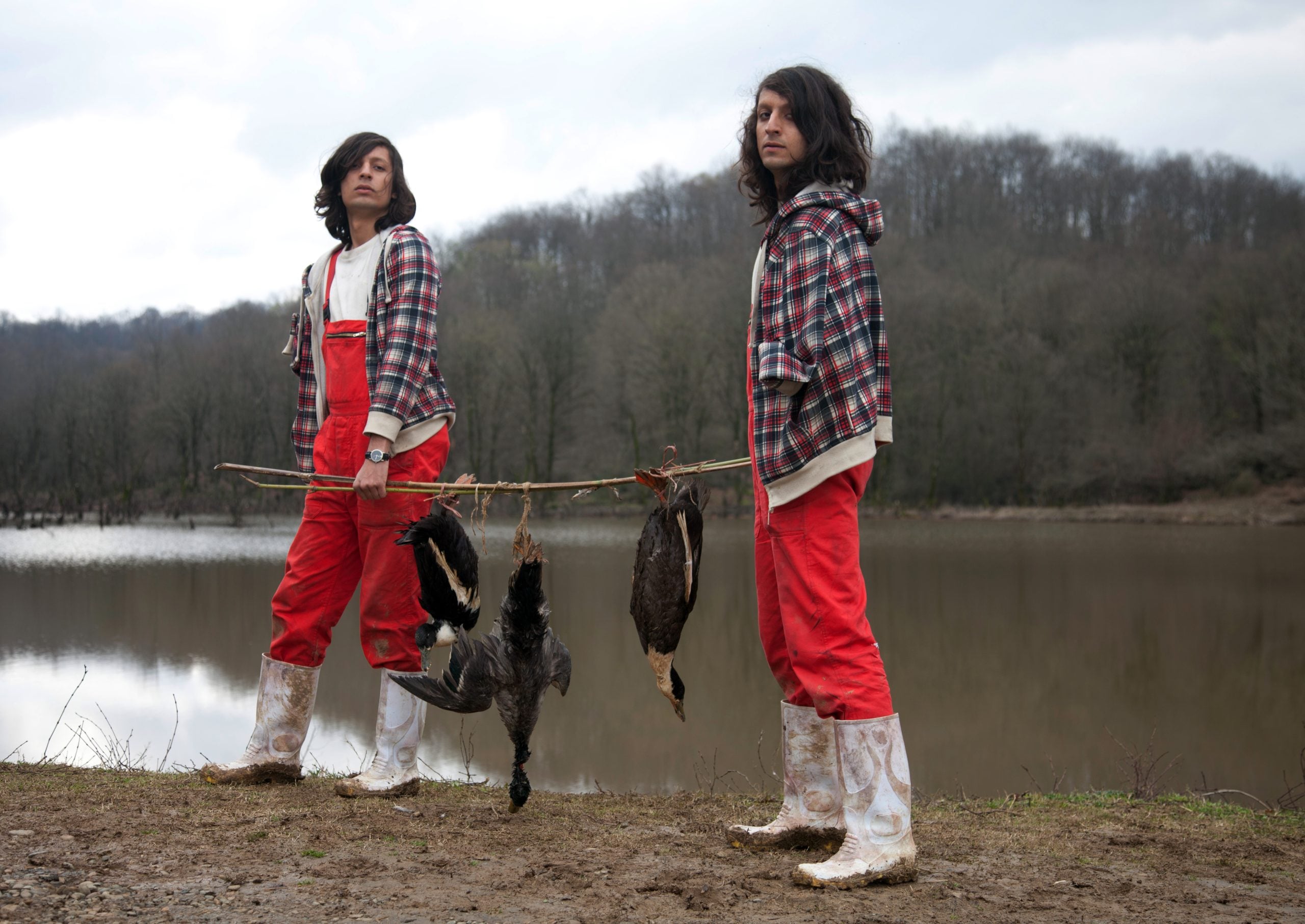 Two people in red overalls and plaid jackets stand by a lake, carrying three large birds on a stick, with a forest in the background.
