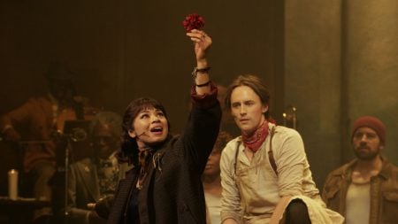 ‘Hadestown: The Musical’ Review – A Magical Capture That Combines The Best of Live Theatre And Film A woman holds up a red flower on stage, with three people seated nearby, in a theatrical performance setting.