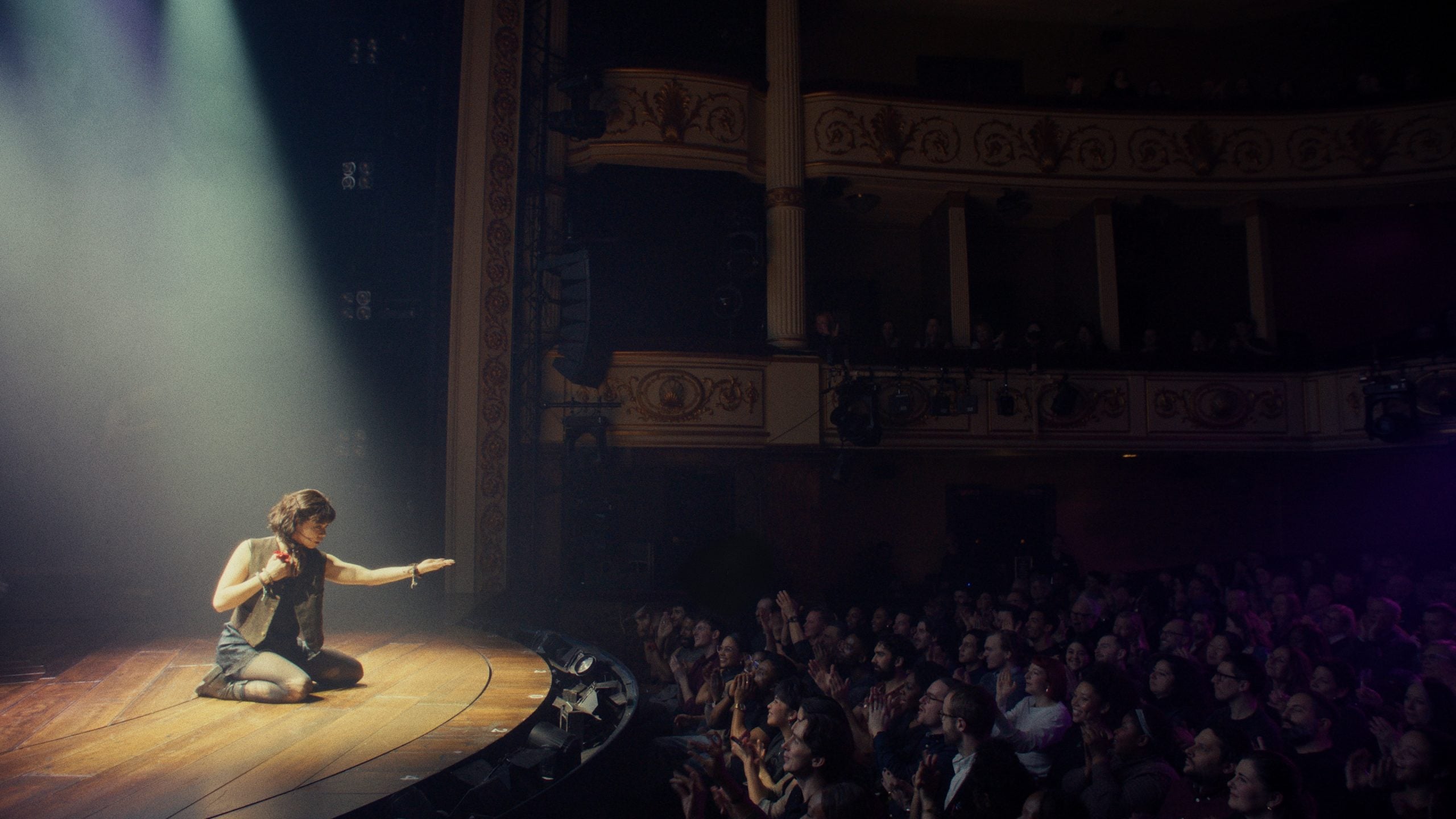 A performer kneels on a stage under a spotlight, reaching towards the audience, while a large crowd watches from their seats in a theater.