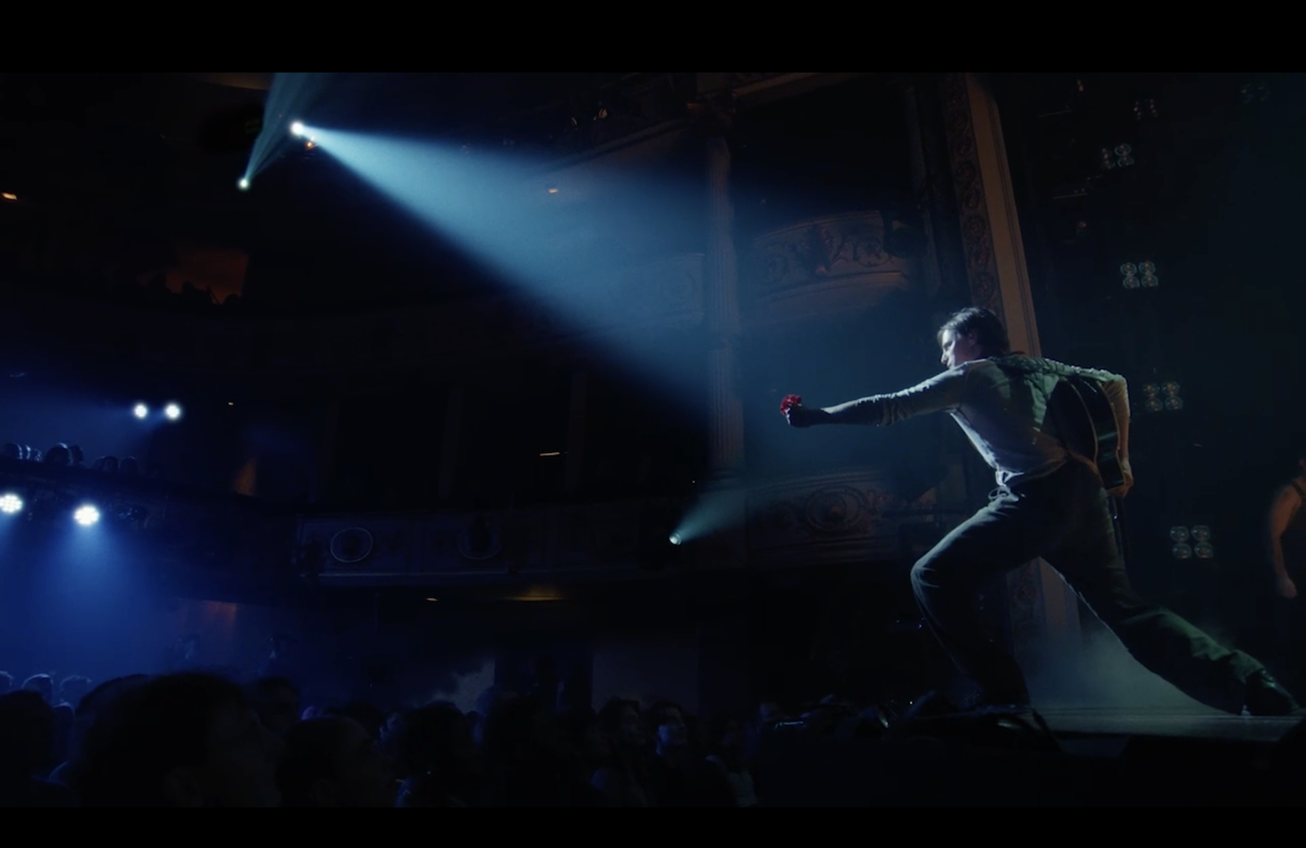 A person stands on stage under a spotlight, extending one arm forward toward an audience in a dark theater.