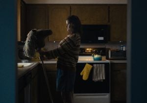 A woman stands in a dimly lit kitchen, holding a toy hobby horse and looking down at it.