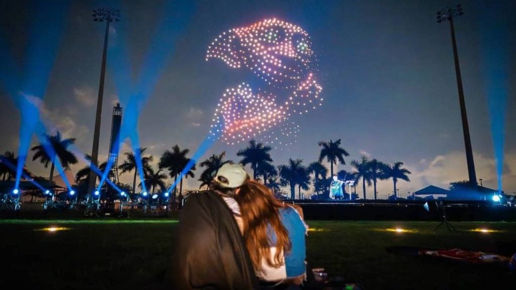 A couple sits on grass at night watching a drone light show forming an image of a dolphin in the sky, with palm trees and stage lights in the background.