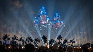 A nighttime outdoor event with a crowd watching a sky display of drones forming a colorful castle shape, with palm trees and stage lights in the foreground.