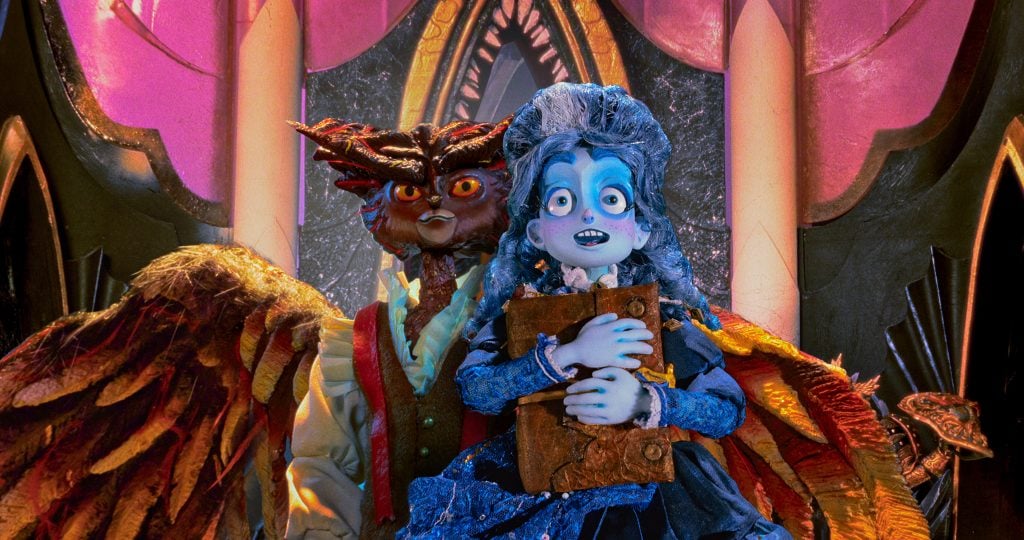 A blue-skinned, wide-eyed character in Victorian attire holds a book, standing in front of a winged, owl-like figure inside a colorful, ornate setting.