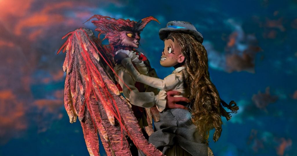 A puppet girl in a gray hat and dress embraces a red-feathered puppet creature against a blurred blue and orange background.