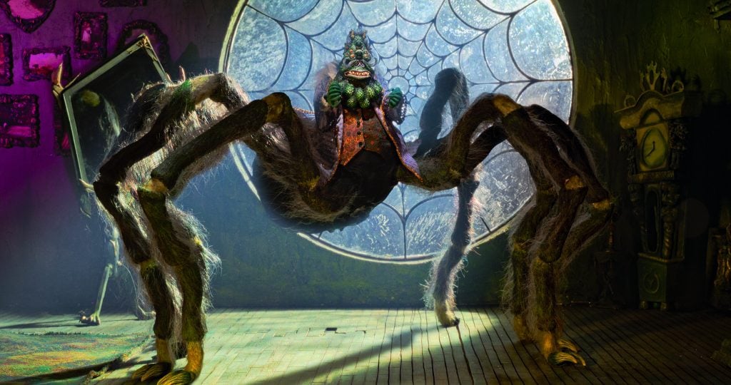A humanoid spider creature wearing a vest stands in front of a large window with a spiderweb pattern in a dimly lit, eerie room.