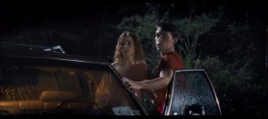 Indican Pictures Debuts Clip From Killer Thriller ‘Jacked’ Ahead of Release (EXCLUSIVE) Two people stand by an open car door at night in a wooded area, looking off-camera with concerned expressions. Rain is visible on the car’s window.