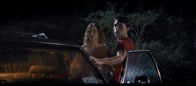 Two people stand by an open car door at night in a wooded area, looking off-camera with concerned expressions. Rain is visible on the car’s window.