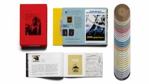 A red box set, an open booklet, posters, documents, and a spread of color swatch discs are arranged on a white background.