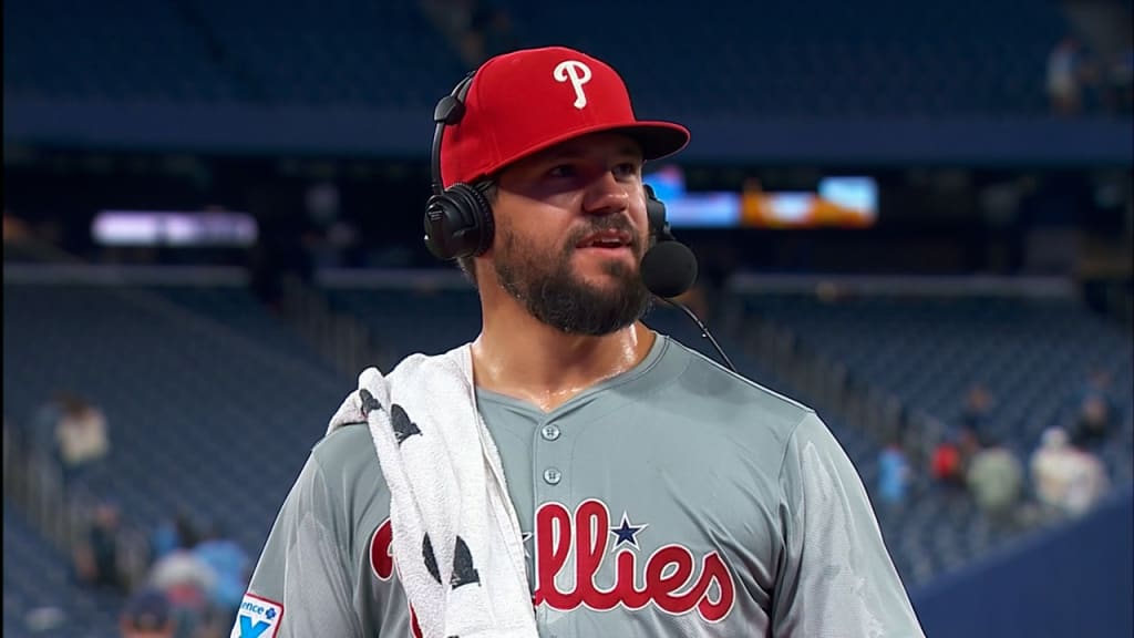 Baseball player in a Phillies uniform and red cap wears a headset and towel over his shoulder during a post-game interview in a stadium.