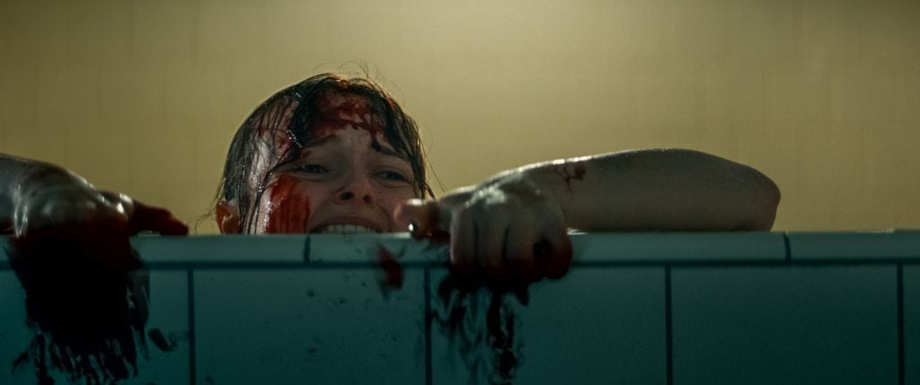 A person with blood on their face and arms grips the edge of a tiled surface, appearing distressed in a dimly lit room.