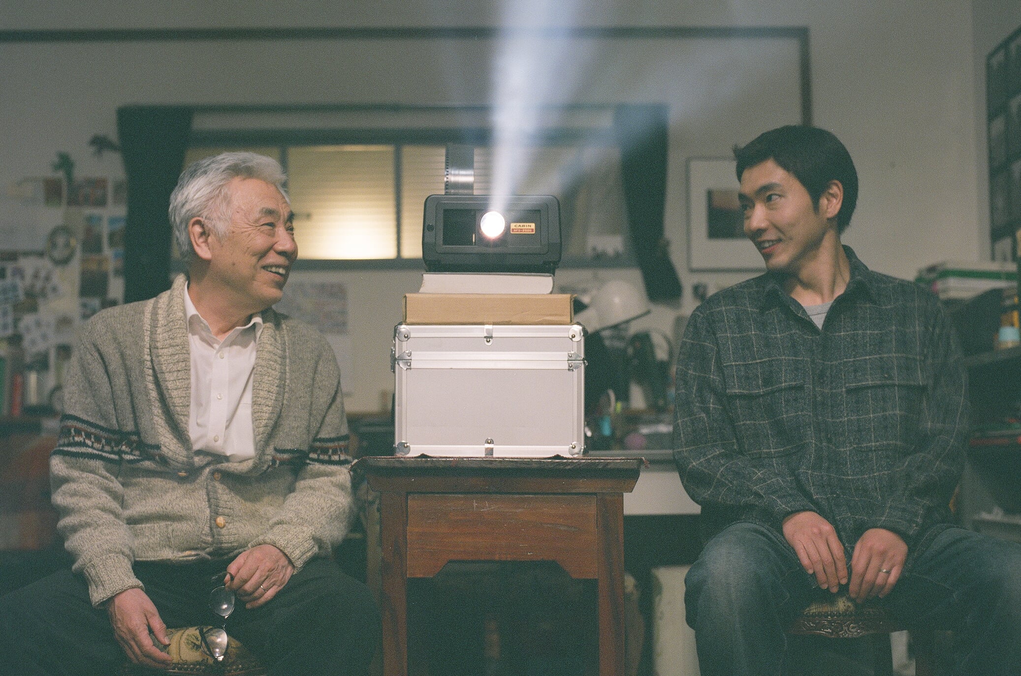Two men sit on either side of a table with a running projector between them, smiling at each other in a warmly lit room.