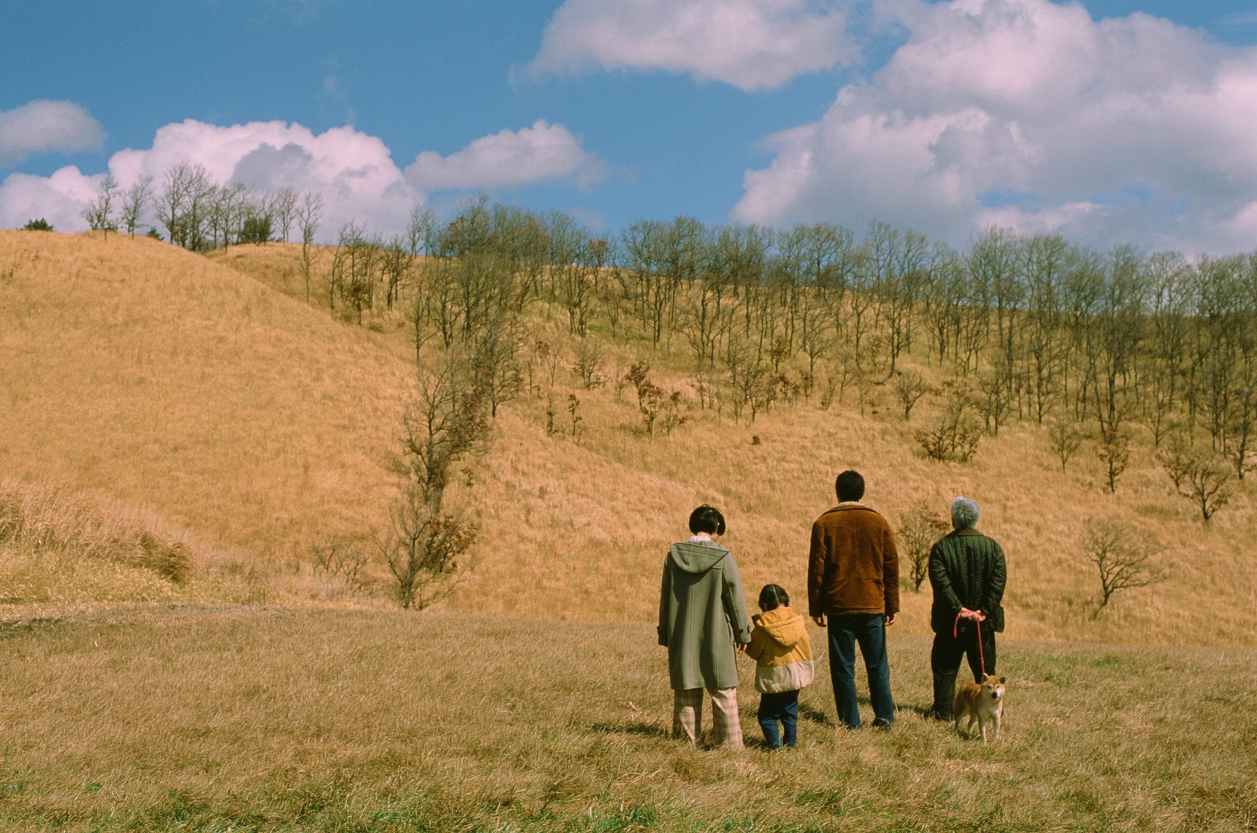 Four people and a dog stand in a grassy field, facing a hill covered with dry grass and scattered trees under a partly cloudy sky.