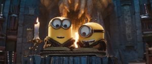 Minions & Monsters Review: A Beautiful Love Letter To B-Movie Cinema Two Minions in robes sit at a stone table reading books by candlelight in a dimly lit, cave-like library setting.
