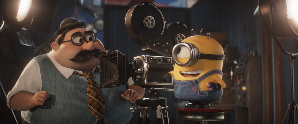An animated character with a mustache stands beside a Minion, who is sitting on a stool and looking through a vintage film camera in a studio setting.
